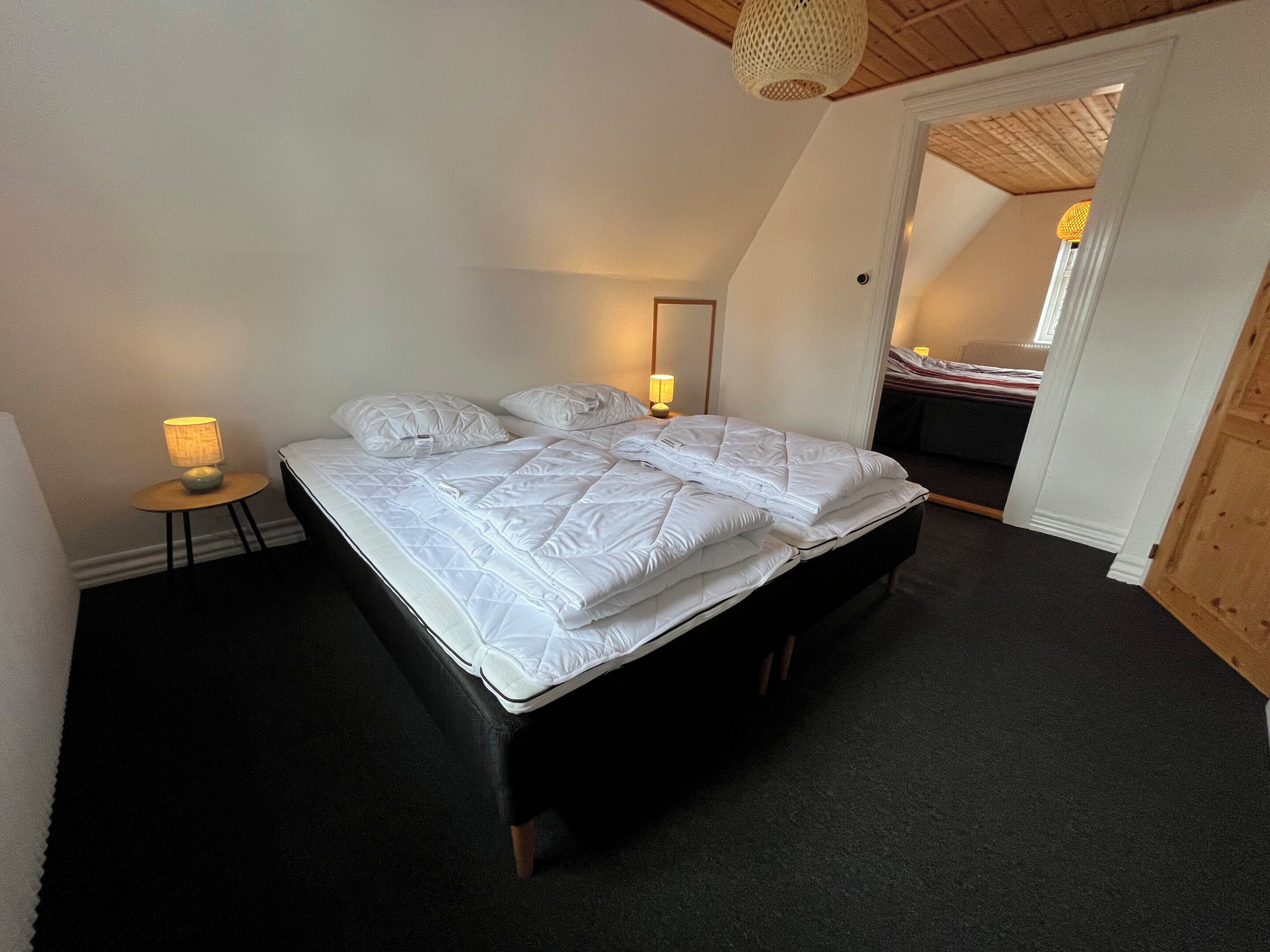 A neatly made bed in a room with a sloped ceiling, a small bedside table with a lamp, and a doorway leading to another room.