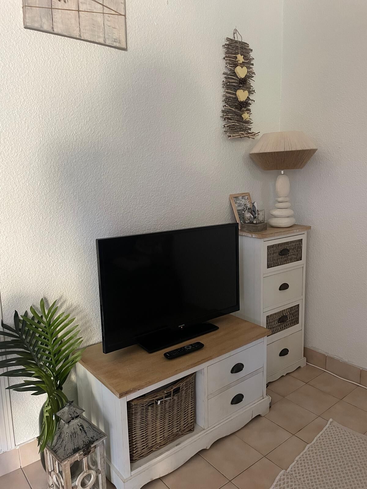 A living room with a flat-screen TV on a wooden stand, a white cabinet with drawers, a wicker basket, a lamp, and a potted plant.