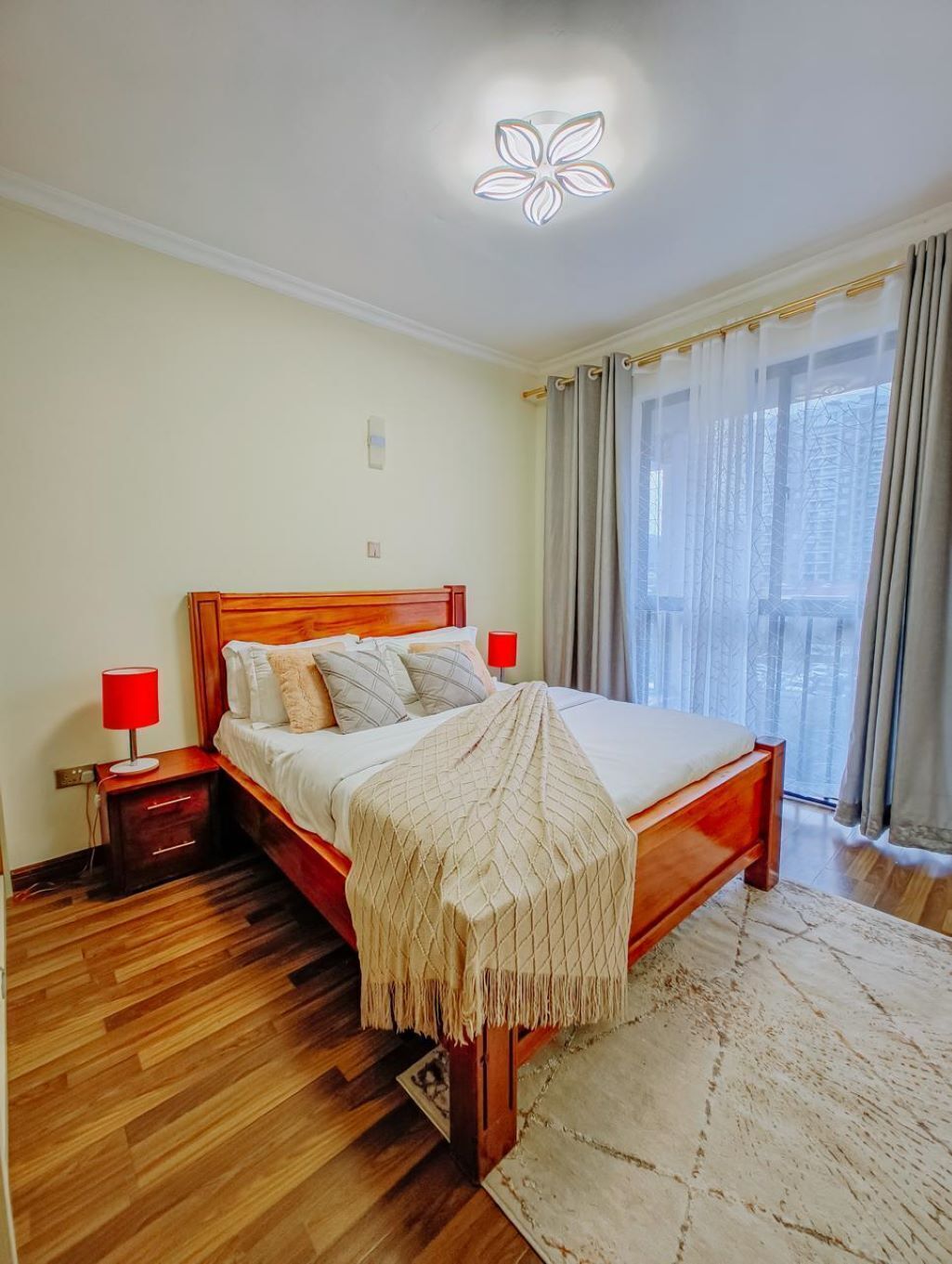 Une chambre à coucher avec un lit en bois, deux tables de chevet, une lampe rouge, un luminaire au plafond et une vue par une fenêtre avec des rideaux.