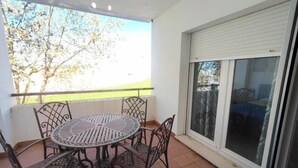 A balcony with a round table and chairs, a window with blinds, and a view of trees. - Aura Conil 32326 in Conil de la Frontera (Conil de la Frontera)