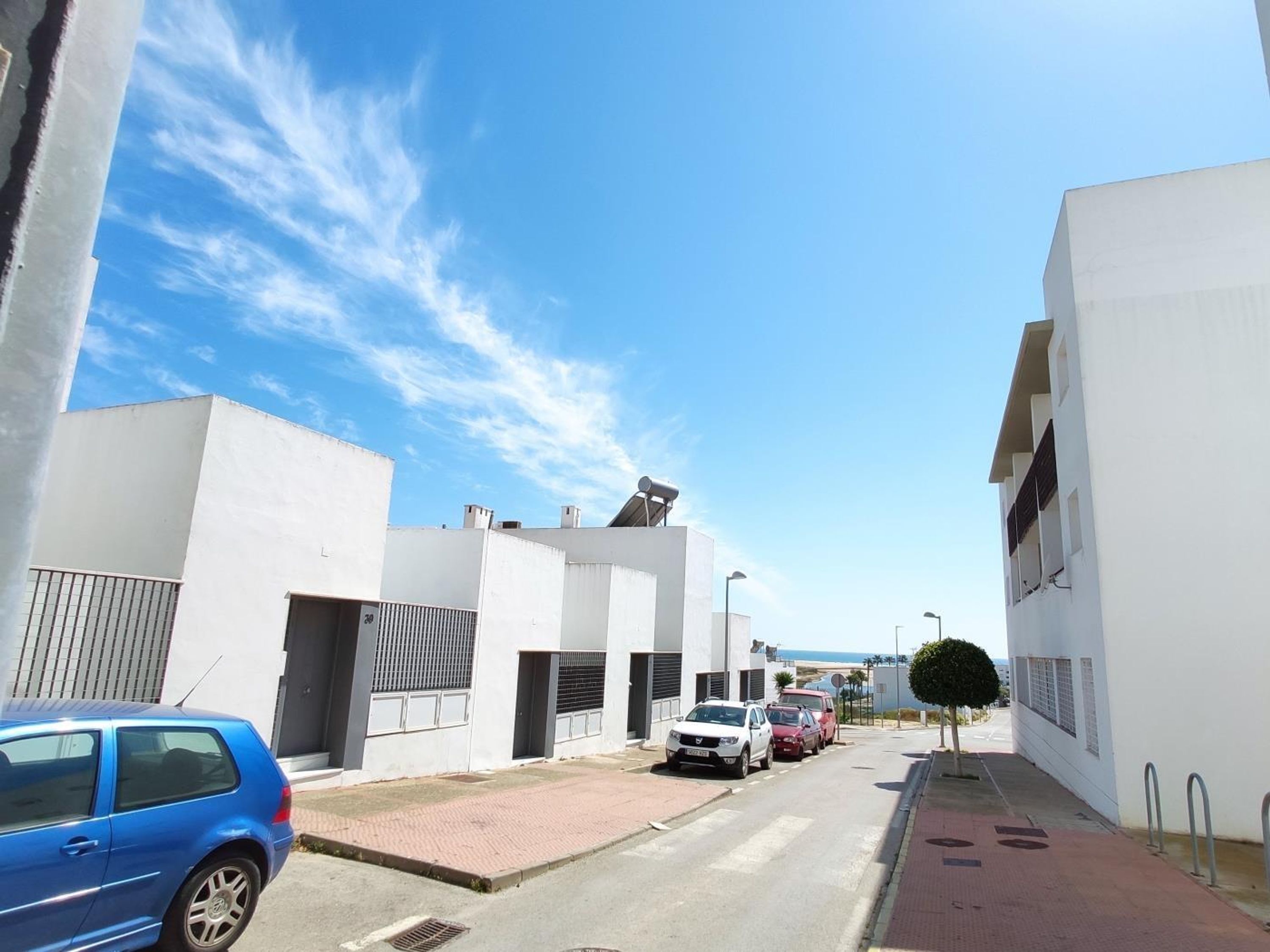 A narrow street with white buildings, parked cars, and a clear sky.