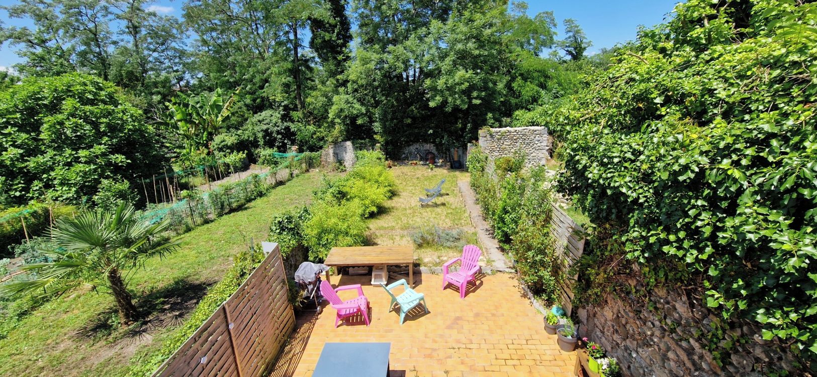 Un espace patio avec une table en bois et deux chaises roses, entouré d’une végétation luxuriante et d’un mur en pierre.