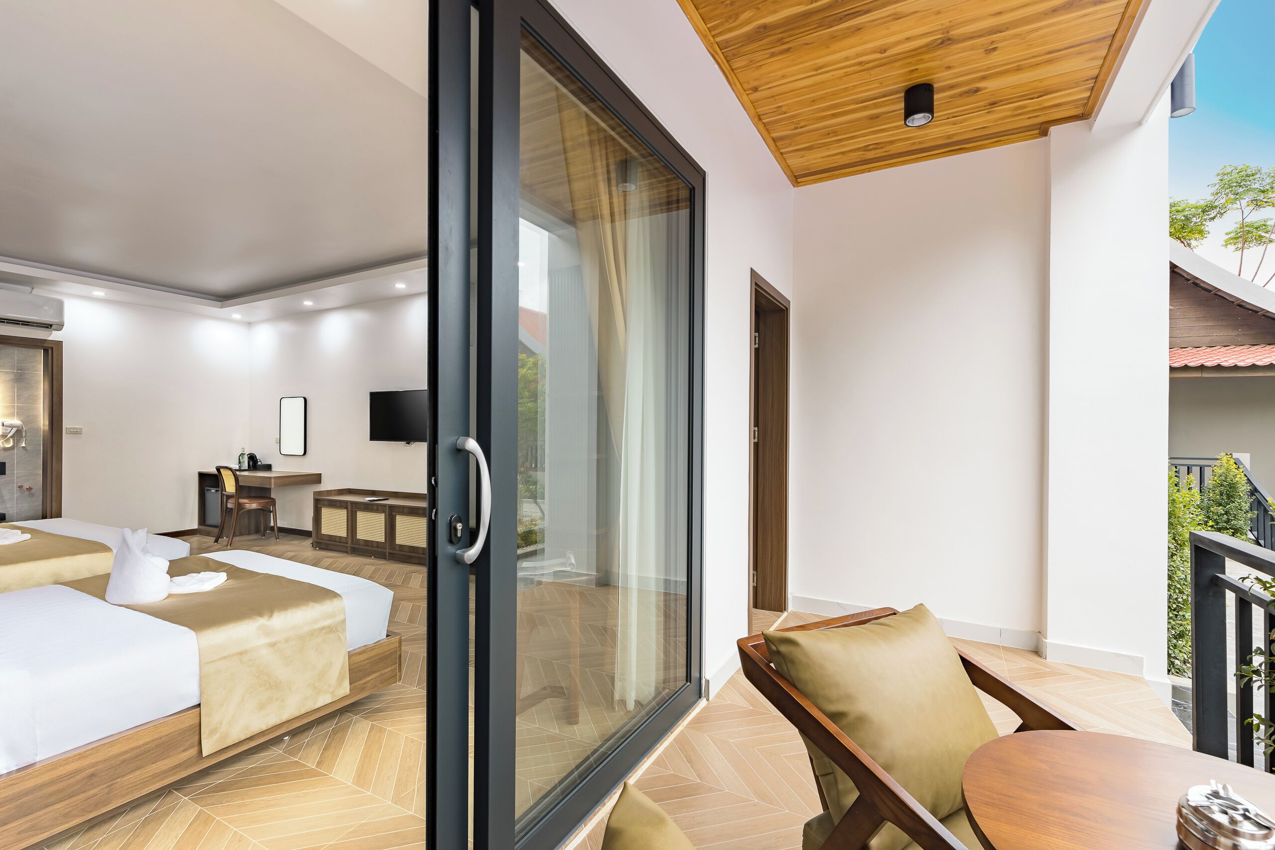 A modern hotel room with a large sliding glass door leading to a balcony, a bed with a wooden headboard, a desk with a chair, and a television on the wall.
