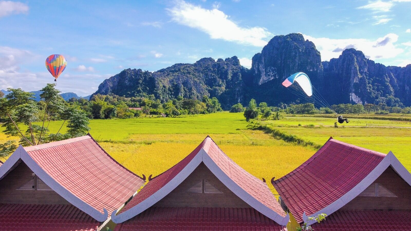 A view of a rural landscape with traditional houses, a hot air balloon, and a paraglider.