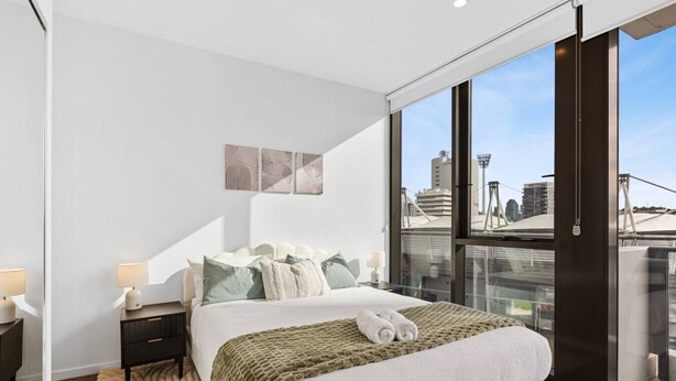 A modern hotel room with a large bed, a nightstand, a lamp, a view of the city through large windows, and a cityscape visible outside. - Walk to The Gabba | Stylish with Rooftop Views (Woolloongabba)