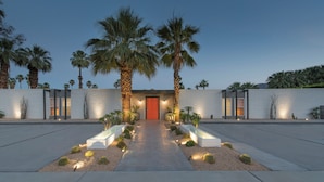 A modern building with a central red door, surrounded by palm trees and a well-lit pathway leading to the entrance.