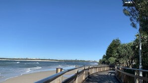 Ein hölzerner Steg verläuft entlang eines Strandes, mit freiem Blick auf das Meer und entfernte Gebäude.