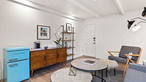 Een moderne woonkamer met een blauwe vintage koelkast, een houten kast, twee ronde tafels, een grijze bank en een deur.
