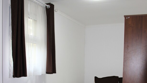 A small, sparsely furnished room with a single bed, a wooden nightstand, a window with curtains, and a patterned carpet. - Sultan Kyzyl-Kiya (Kyzyl-Kiya)