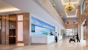 A modern hotel lobby with a reception desk, a large mural of a coastal scene, a chandelier, and a luggage cart. - Vienna Sleep International Hotel (Shenzhen)