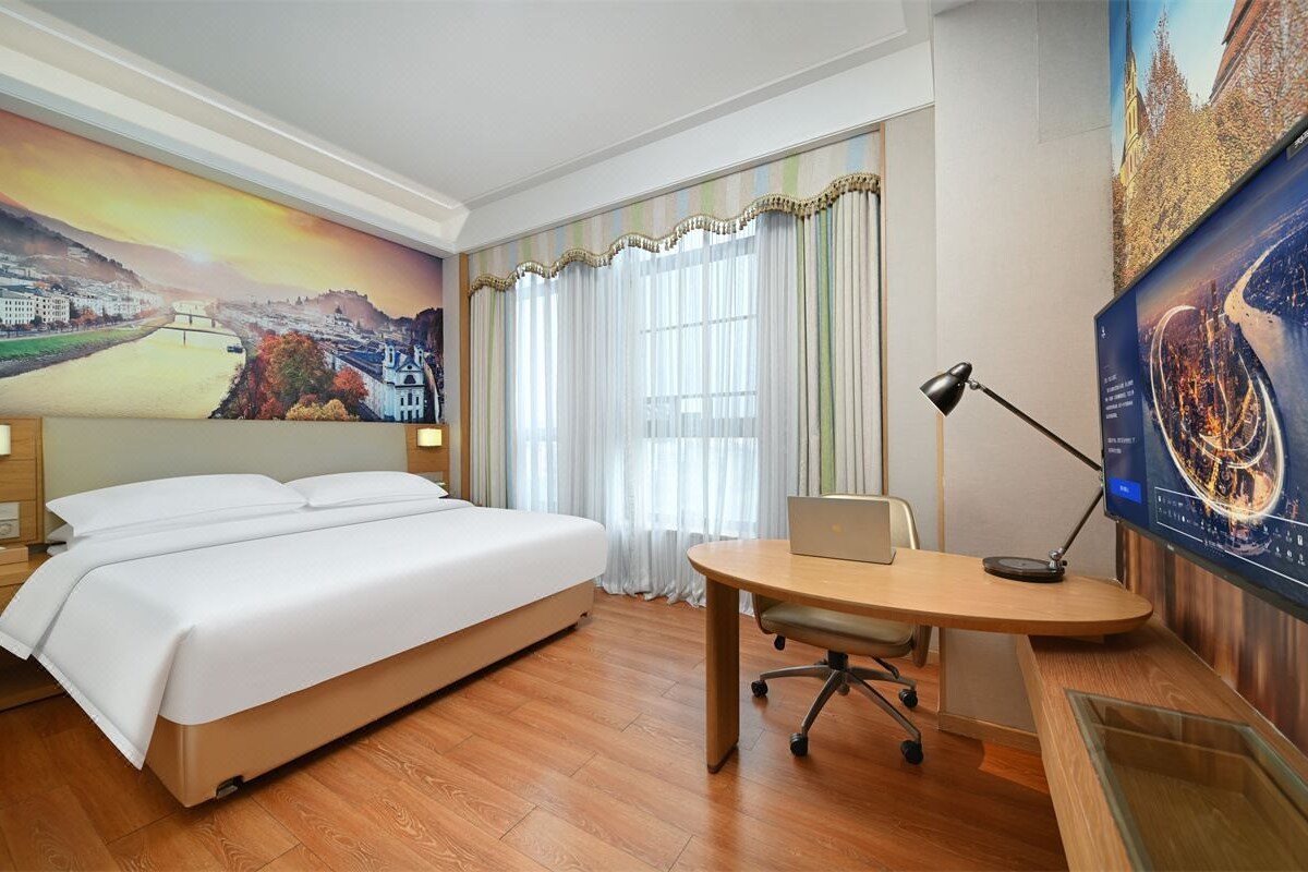 A hotel room with a bed, a desk with a laptop, a TV, and a large window with curtains.