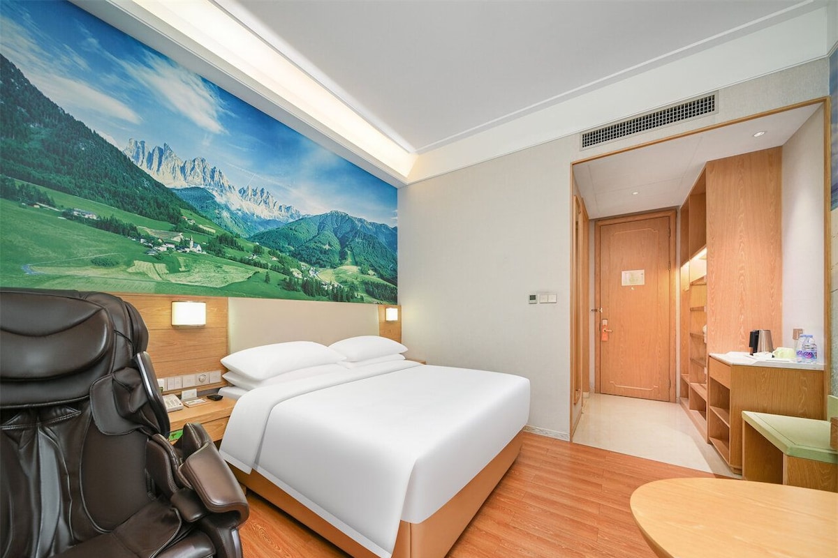 A hotel room with a bed, a massage chair, a desk, and a view of a mountain landscape.