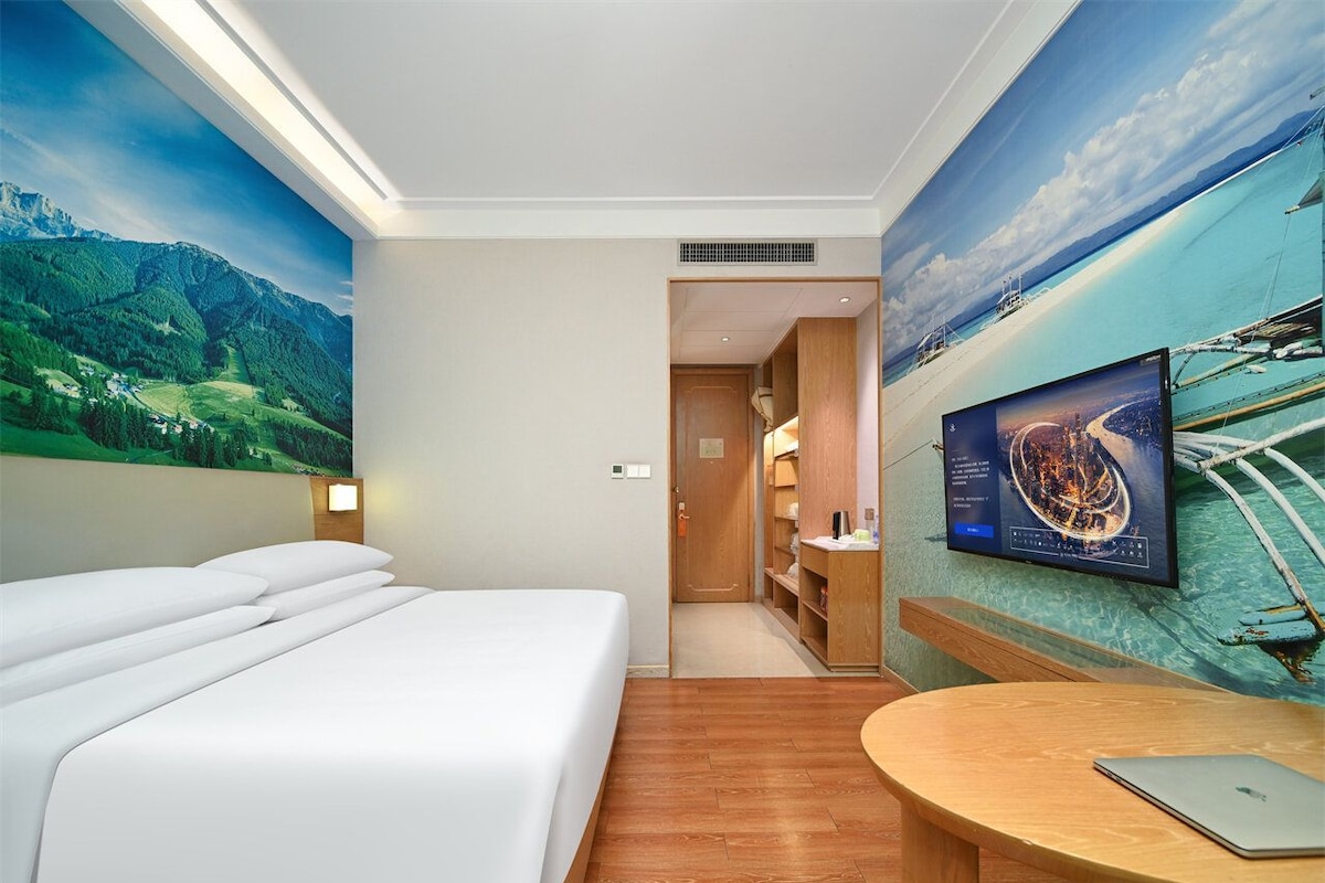A hotel room with a bed, a desk, a TV, and a mural of a landscape.
