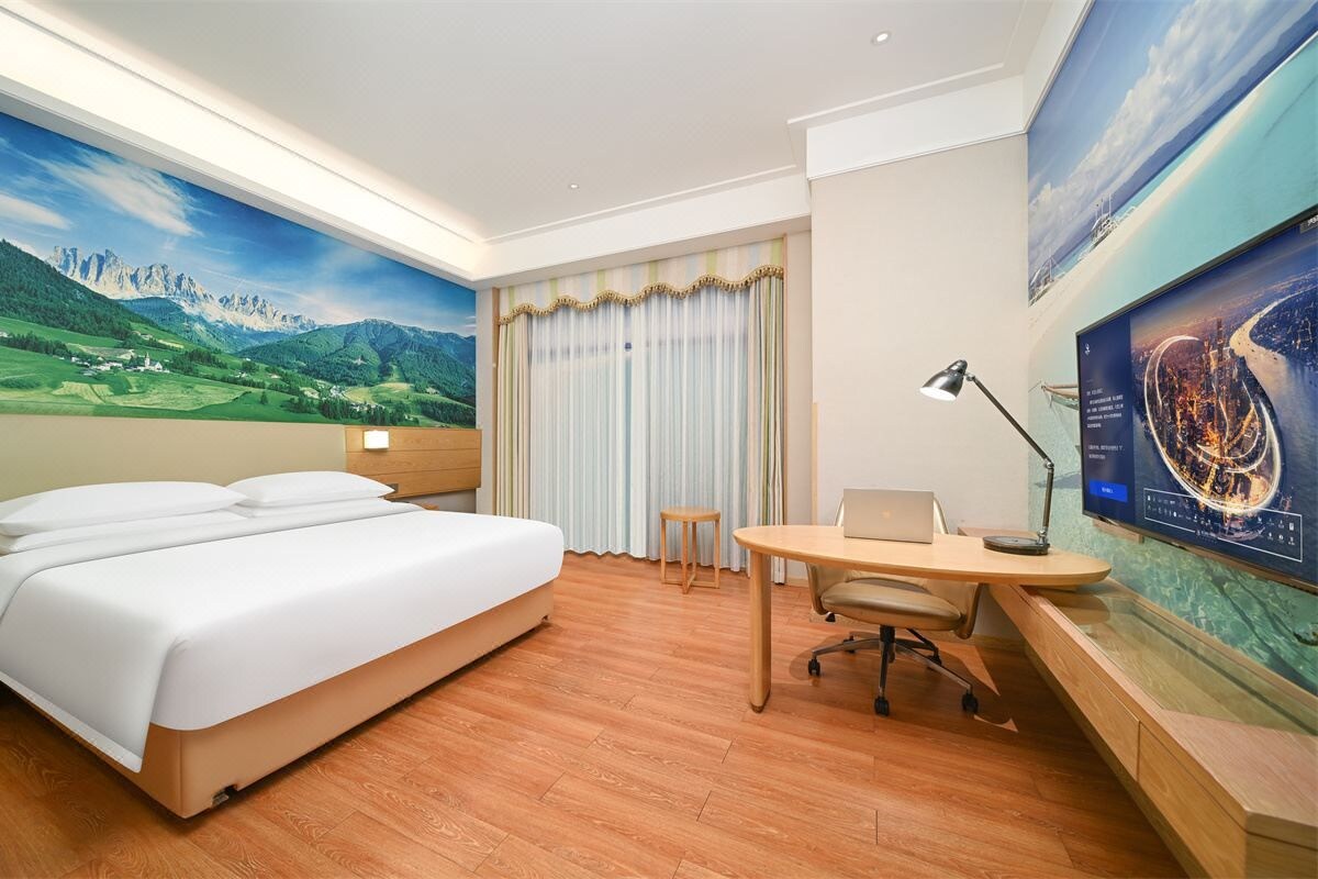 A hotel room with a bed, desk, chair, and a large window with curtains.