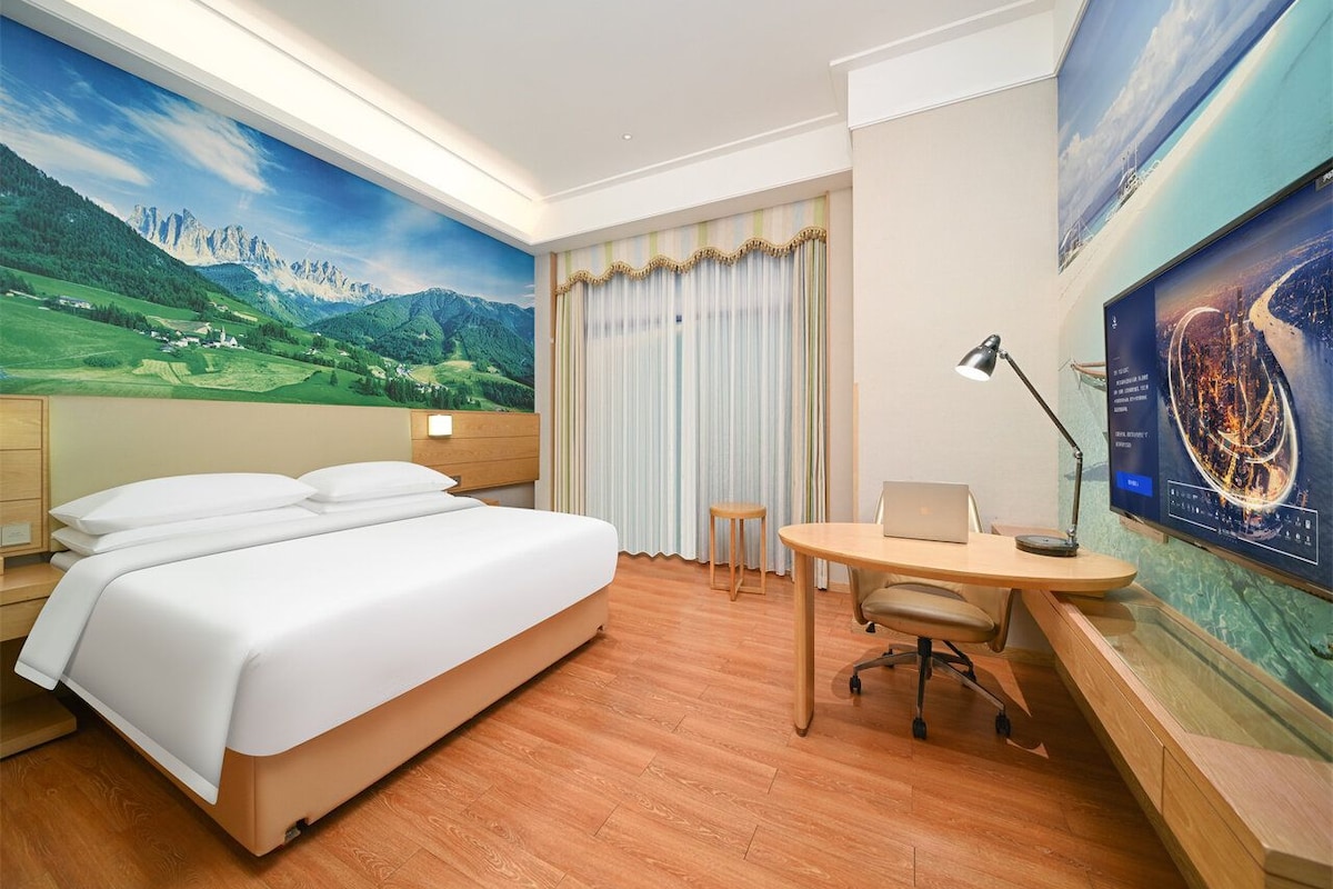 A hotel room with a bed, desk, chair, and large windows with curtains.