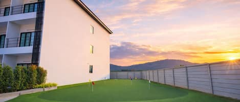 A modern building with a flat roof and a flat facade, a well-maintained lawn with a putting green, and a clear sky with the sun setting in the background.