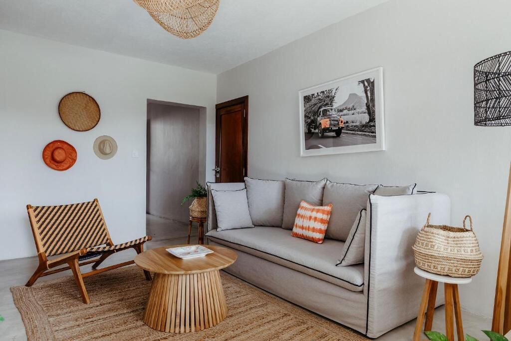 A modern living room with a sofa, a wooden coffee table, and a chair. There is a door leading to another room, a framed picture on the wall, and a woven basket on a small table.
