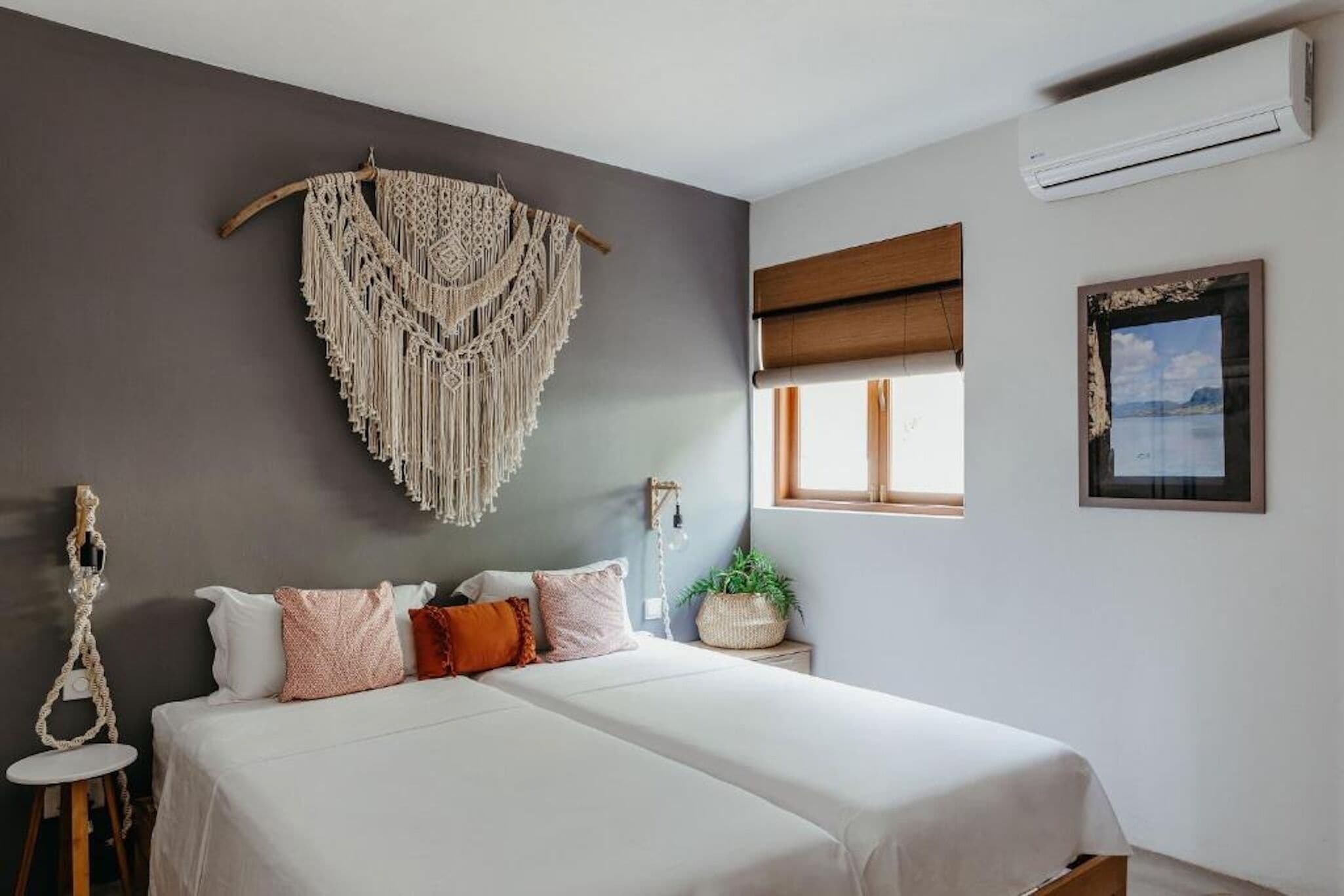 A bedroom with a bed, macrame wall hanging, a window with a view, and an air conditioning unit.