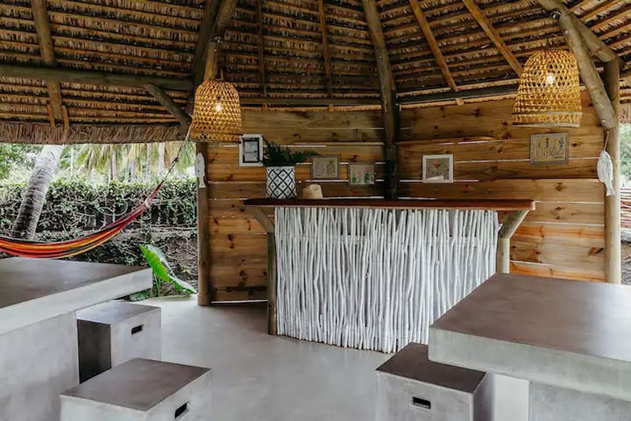 Una estructura de madera con techo de paja, una zona de bar con mostrador tejido y una hamaca afuera.