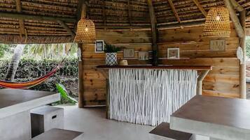 Una estructura de madera con techo de paja, una zona de bar con mostrador tejido y una hamaca afuera.