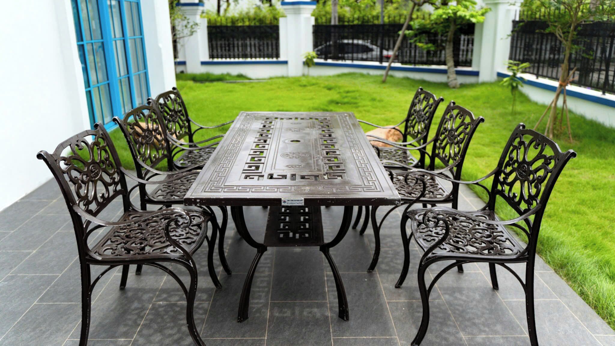 A rectangular outdoor table with intricate metalwork, surrounded by six chairs with similar design, set on a tiled patio.