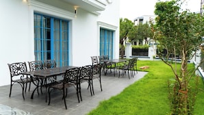 A patio with a table and chairs, a white building with blue-framed windows, and a green lawn. - Soleil Villa (Bai Chay)