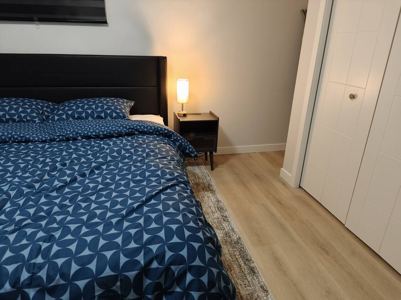 Quarto com cama, mesa de cabeceira, luminária e porta.