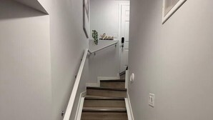 A staircase with wooden steps and white handrails, leading to a door with a light switch and a vent on the wall. - Modern 1Bedroom Stylish Private Basement Suite (Leduc)