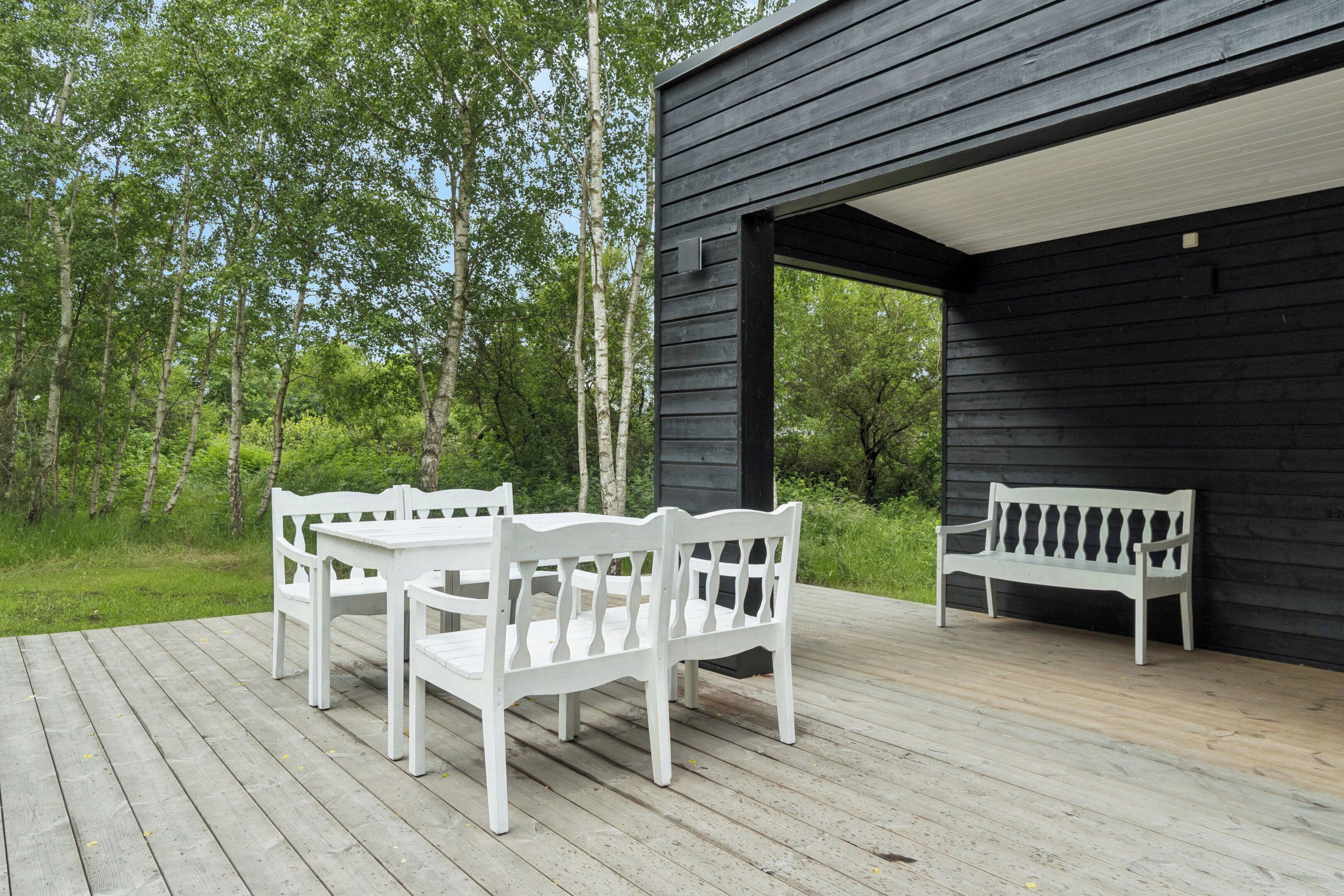 A wooden deck with white outdoor furniture, a black building, and a forested background.