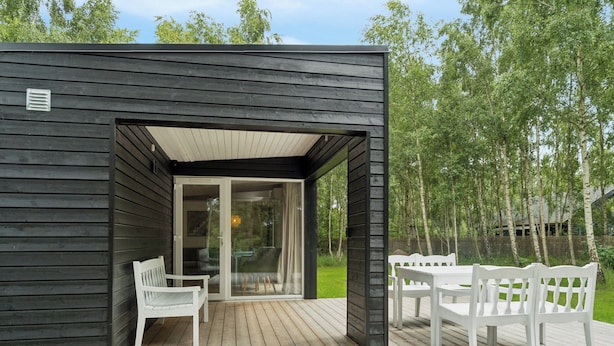 A wooden deck with a covered seating area, a glass door entrance, and a view of a green landscape. - Cozy retreat near the Djursland coast (Ørsted)