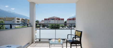 A balcony with a chair and a table, overlooking a parking area and buildings with a mountain in the background.