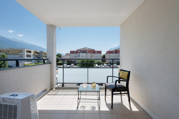 Un balcón con una silla y una mesa, con vistas a un estacionamiento y edificios, con una montaña al fondo.