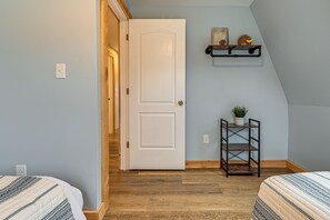 Une chambre avec une porte blanche, un plancher en bois, un lit recouvert d’une couvre-lit à motifs, une petite étagère ornée d’objets décoratifs et une plante en pot.