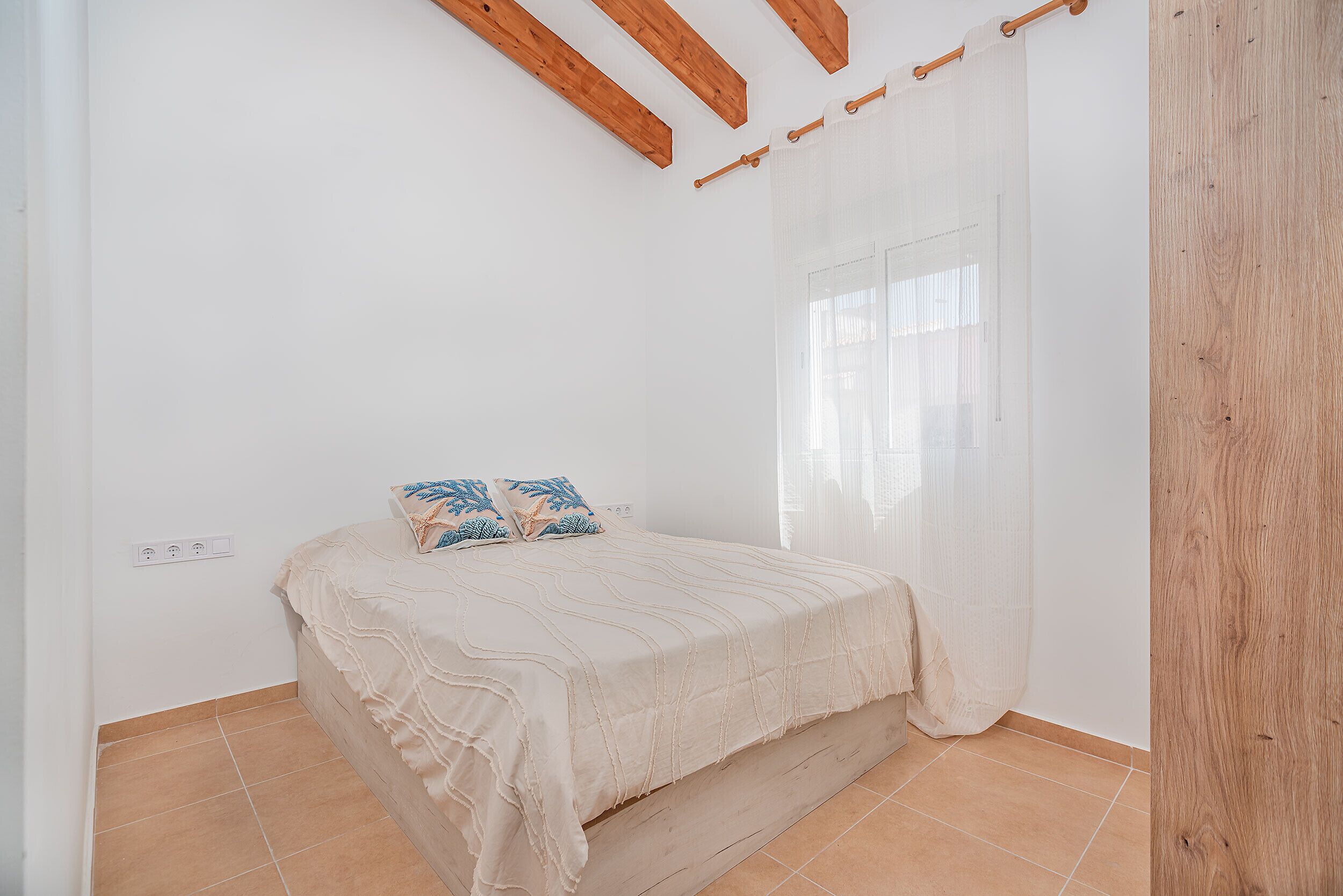 A bedroom with a bed, wooden beams, a window with sheer curtains, and a tiled floor.