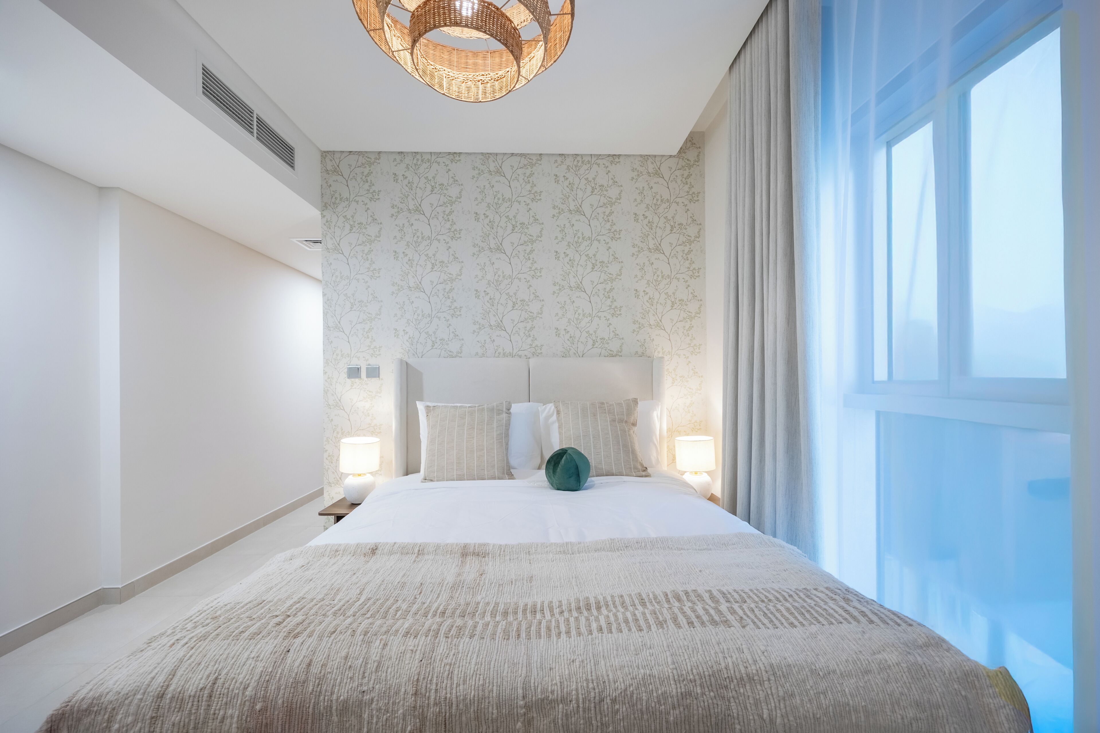 A modern bedroom with a large bed, a headboard, bedside lamps, and a view of the outdoors.