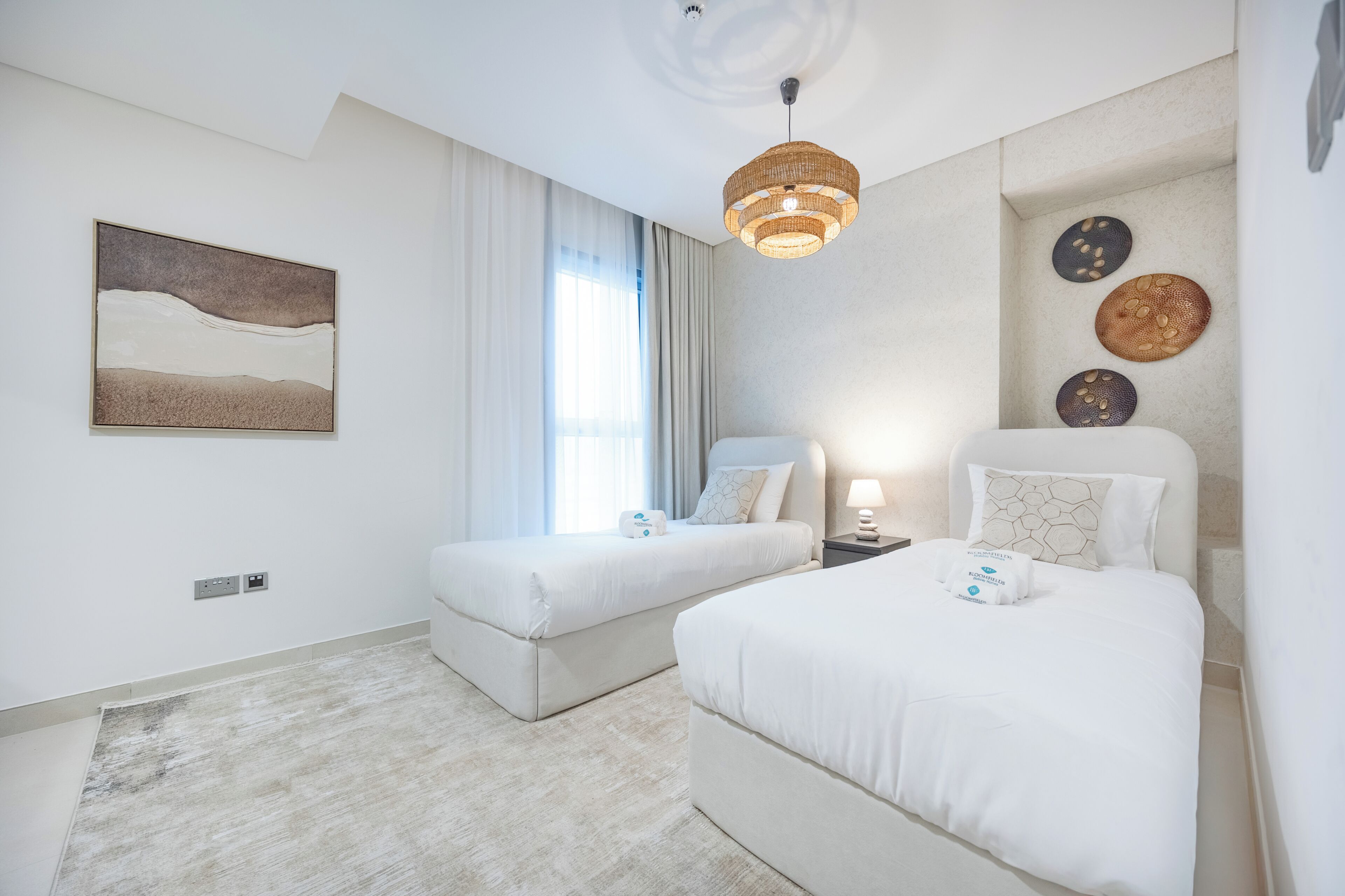 Quarto de hotel com duas camas, uma janela ampla com cortinas, uma obra de arte na parede e um tapete texturizado.
