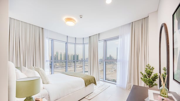 Une chambre d’hôtel moderne avec un grand lit, une table de chevet avec une lampe, une vue sur le paysage urbain grâce à de grandes fenêtres, et un bureau aux tons sombres agrémenté d’un vase de fleurs.