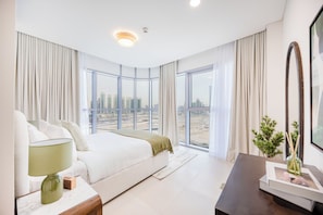 Ein modernes Hotelzimmer mit einem großen Bett, einem Nachttisch mit Lampe, Blick auf die Stadt durch große Fenster und einem dunklen Schreibtisch mit Blumenvase.