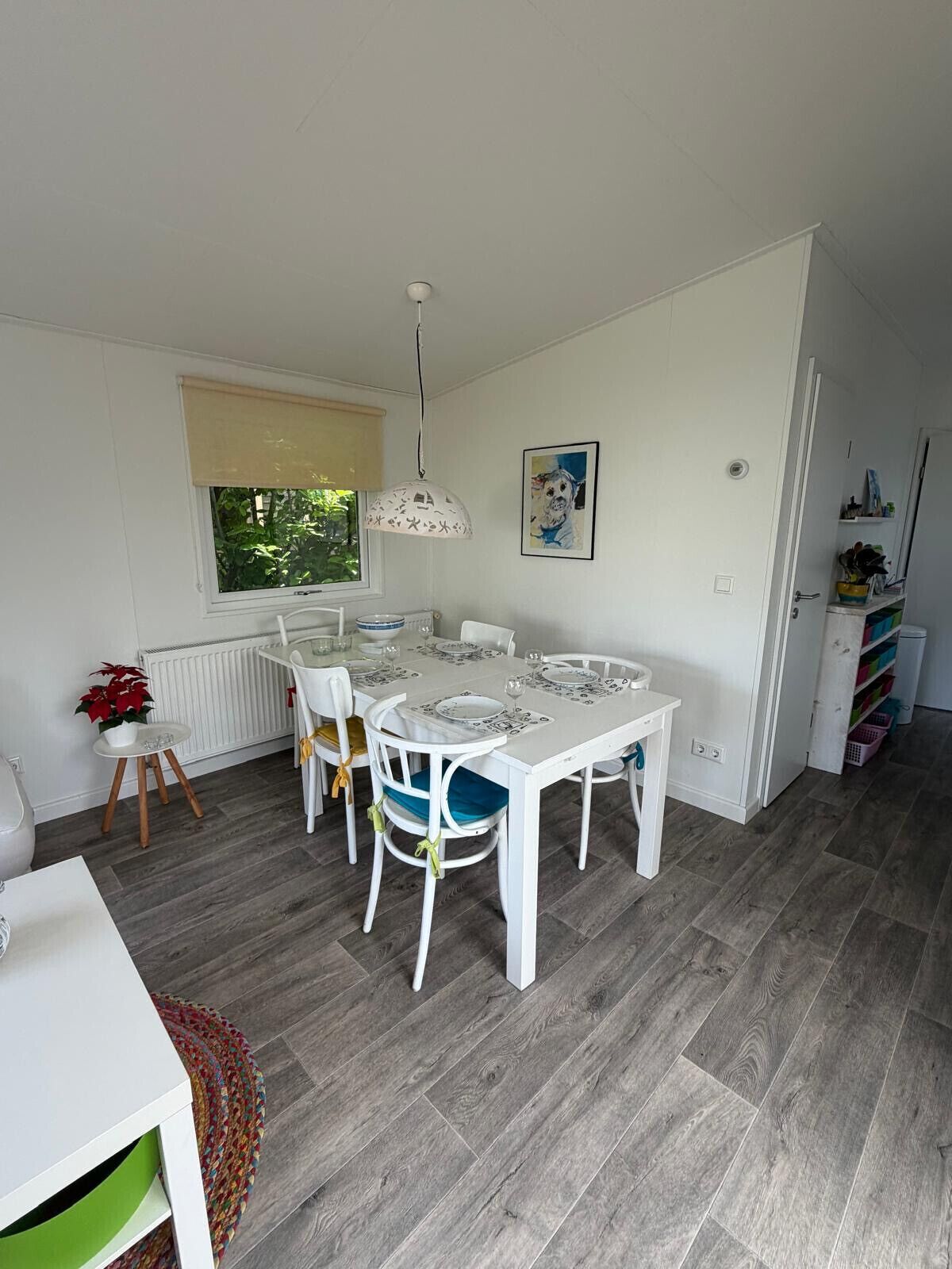 A dining area with a white table and chairs, a radiator, a window with a beige blind, and a hanging light fixture.