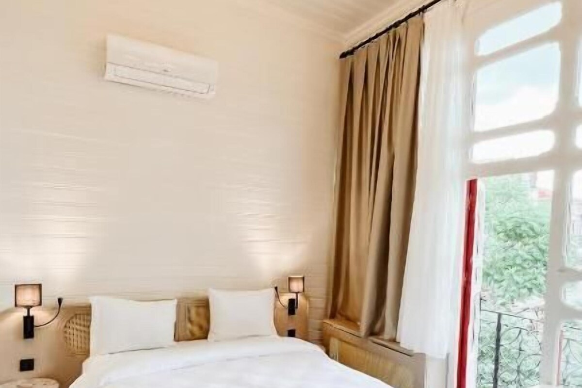A bedroom with a large bed, bedside lamps, a chandelier, a window with curtains, and a view of greenery outside.