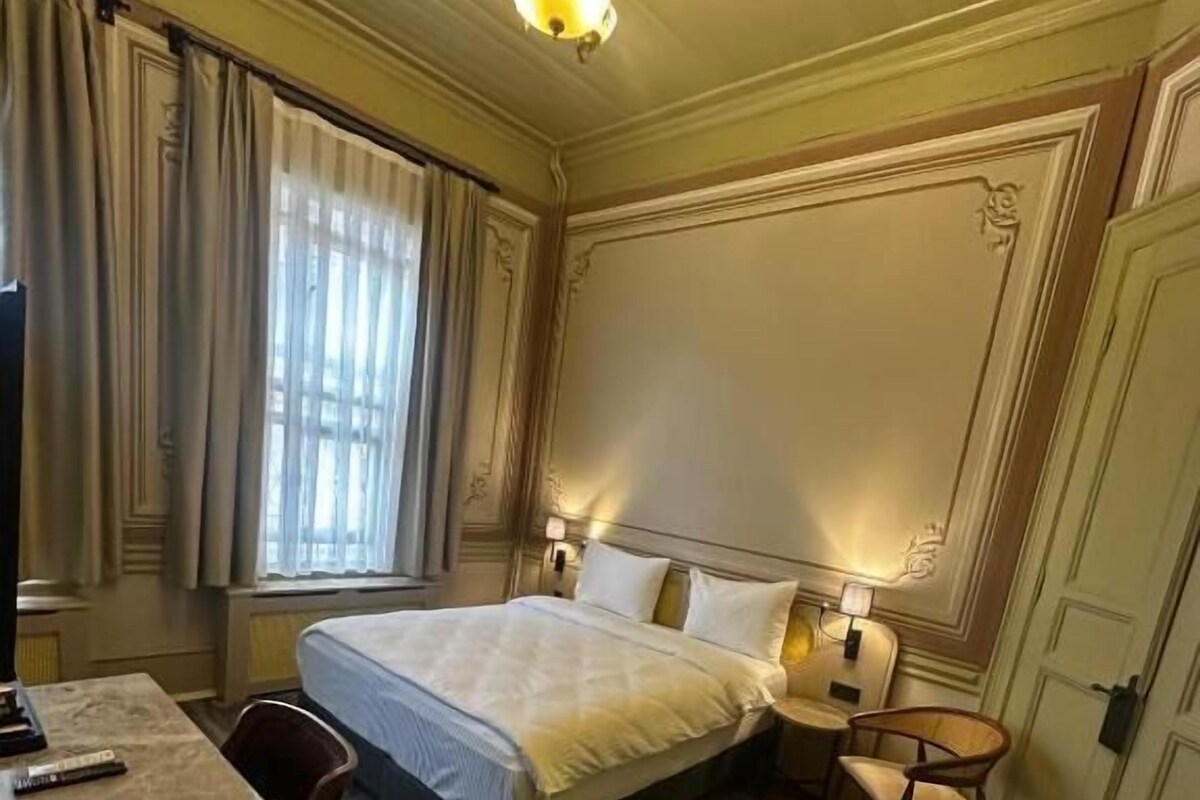 A hotel room with a bed, a desk, a chair, a lamp, and a chandelier.