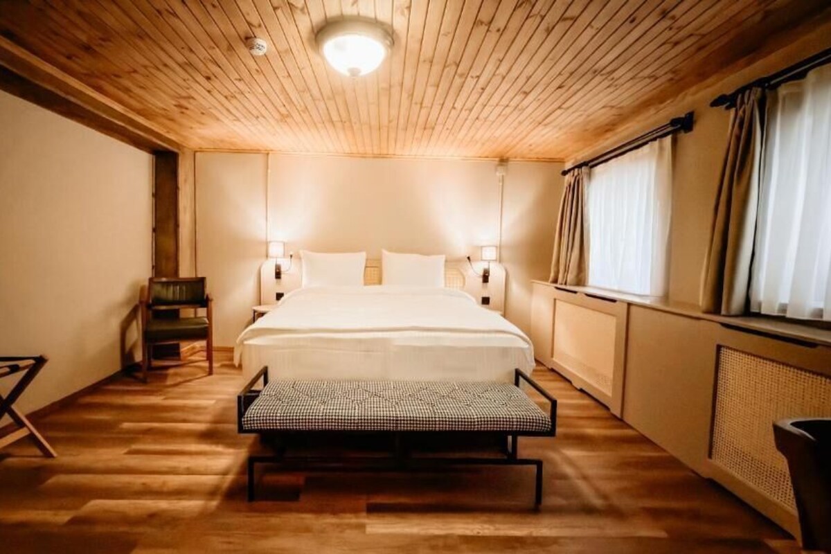 A wooden-paneled room with a bed, bedside lamps, a bench, and a radiator.