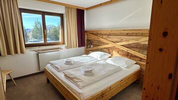 Een slaapkamer met een houten bed, een raam met gordijnen, een bureau en uitzicht op bomen en bergen.