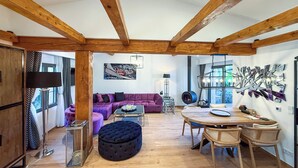 Een ruime kamer met houten balken, een paarse bank, een ronde tafel en een hanglamp.
