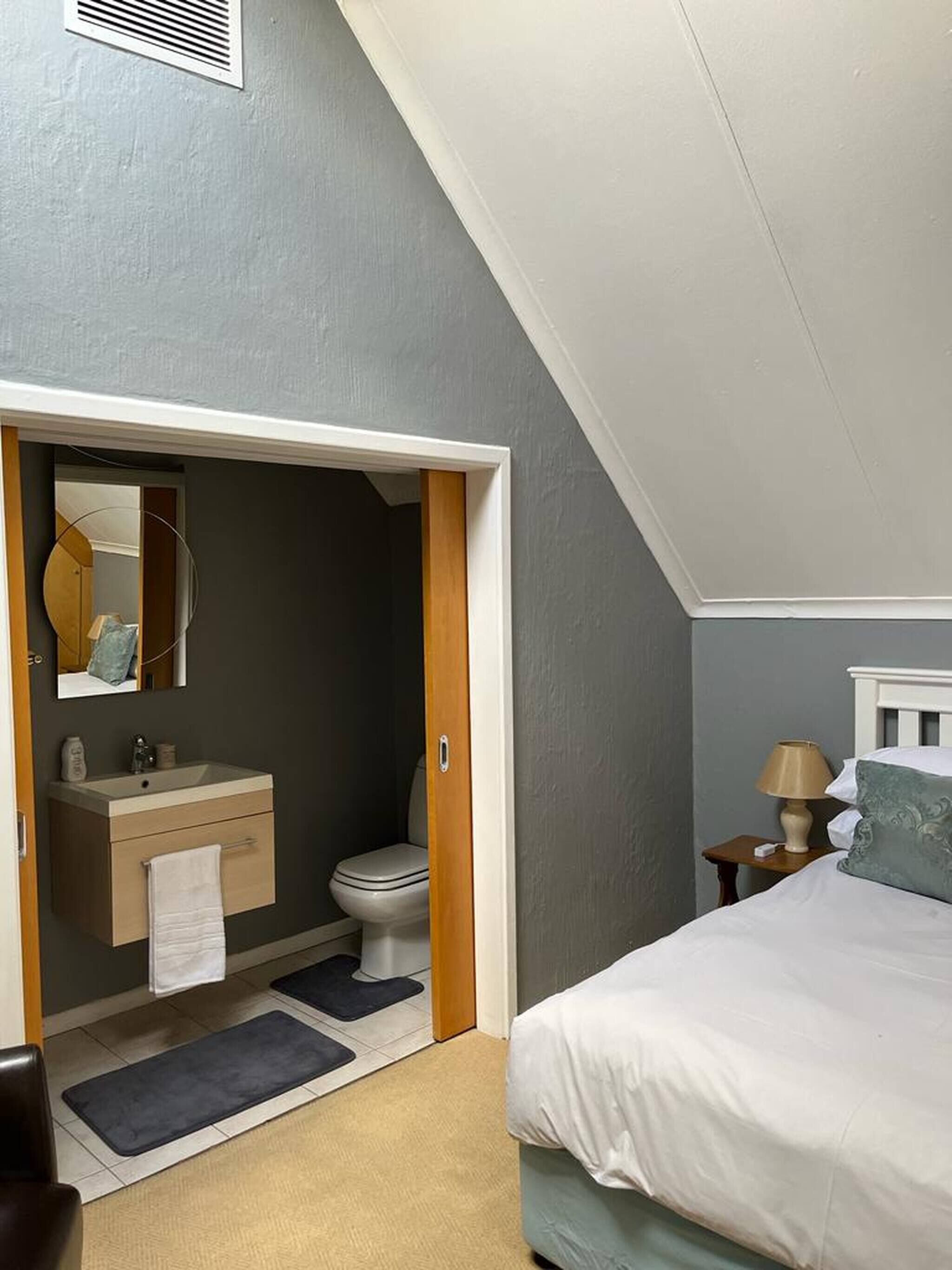 Una camera da letto con un letto, un comodino con una lampada, un bagno con lavandino e WC, e uno specchio.