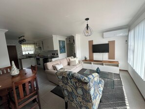 Una sala moderna con un sofá, un sillón estampado, una mesa de comedor y una cocina al fondo.
