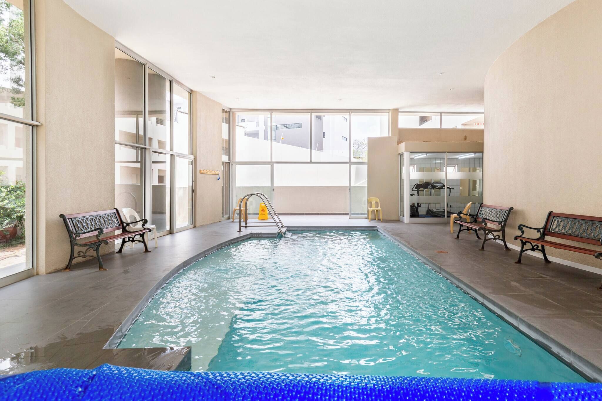 Piscina interna com bancos, portas de vidro e vista para prédios lá fora.
