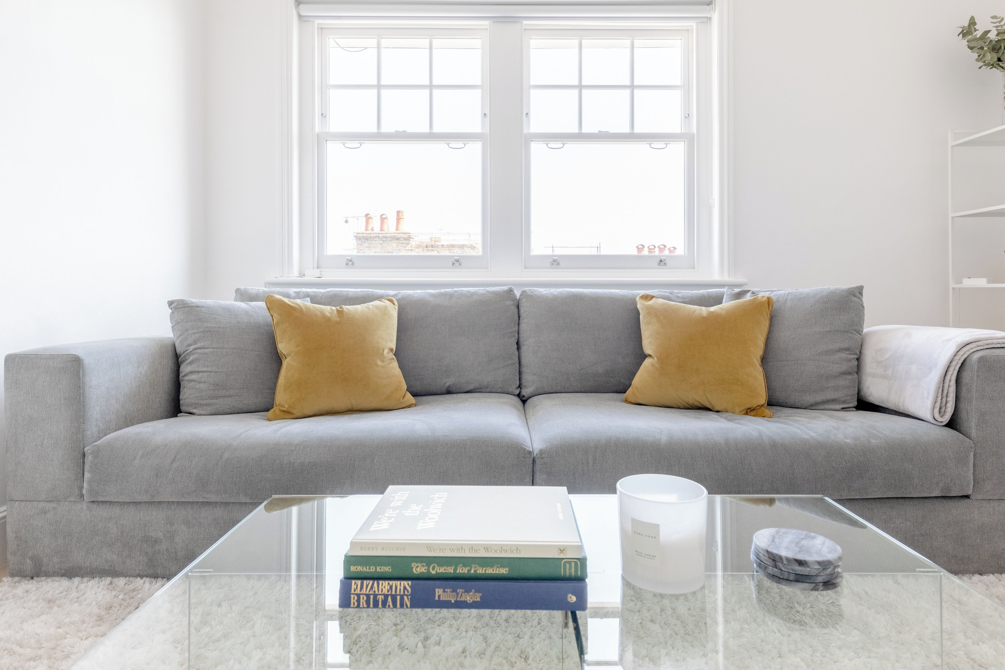 A modern living room with a grey sofa, two mustard-colored cushions, a glass coffee table with books and a candle, and a view of buildings through large windows.
