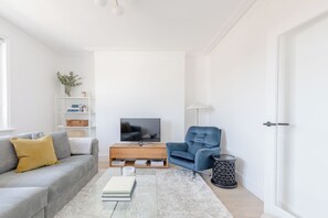 A modern living room with a grey sofa, a blue armchair, a flat-screen TV, a bookshelf, and a small side table. - Sleek Scandi Inspired 2BDR Flat inFulham (London)
