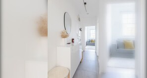 Ein heller Flur mit weißer Wand, einem Spiegel und einer Vase mit getrockneten Blumen. Im Hintergrund ist eine Couch mit einem gelben Kissen zu sehen.
